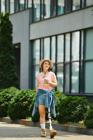 A young woman with a prosthetic leg walks through a city park, holding a cup of coffee and smiling.の写真素材