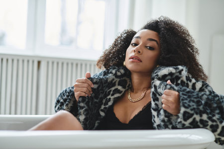 A woman with an afro in a fashionable leopard-print coat sits in a white bathtub and looks directly at the camera.の写真素材