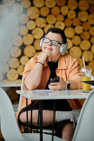 A man with Down syndrome sits in a cafe, headphones on and a phone in his hand, enjoying the day.の写真素材