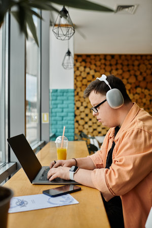 A man with Down syndrome works on a laptop in a cafe, headphones on, with a glass of juice nearby.の写真素材