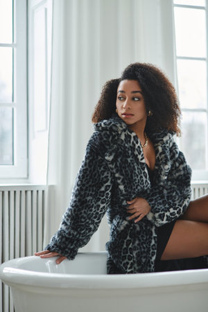 A fashionable African American woman in a trendy leopard print coat poses in a studio setting, leaning against a white bathtub.の写真素材