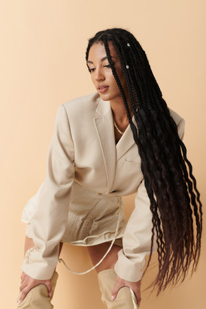 A young woman with long black braids poses in a beige blazer, skirt, and thigh-high boots.の写真素材