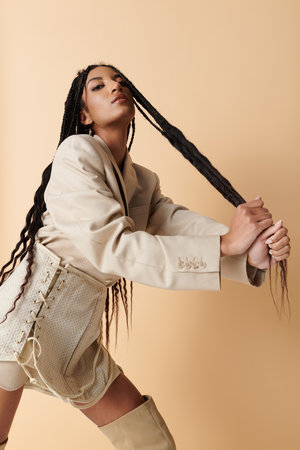 A stylish young woman with long braids poses in a beige blazer and skirt, her hand pulling her braids back.の写真素材