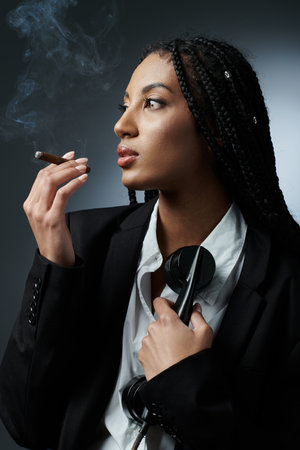 A chic African-American woman in her 20s with braids, white shirt, and black blazer, poses against a gray background smoking a cigarの写真素材