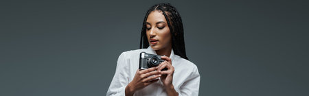 A young woman with braided hair poses against a grey background, holding a camera in her hands.