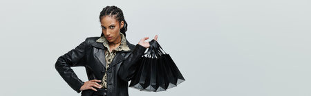 A confident young African American woman in black leather and animal print, holding black shopping bags against a grey backdropの写真素材