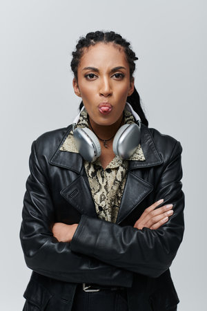 A young woman with braids and a playful expression poses in a black leather jacket with a snake print shirt and silver headphones.の写真素材