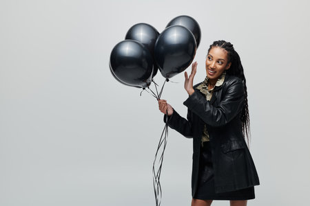 A young woman wearing a black leather jacket and skirt holds a bunch of black balloons while posing against a grey backdrop.の写真素材