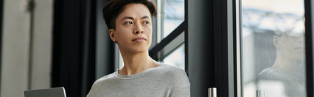 A young man, sitting in a modern office, gazes out the window, contemplating his startup dreams.