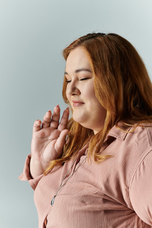 A plus size woman in a pink shirt, with her eyes closed, raises her hand as if in prayer.