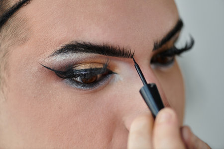 A queer person applies eyeliner to their upper eyelid.