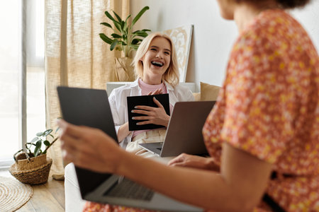 Two women, one with blonde hair, laugh while working on laptops in a home office.