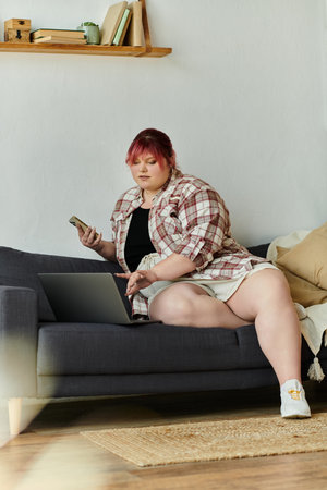 A plus size woman in a plaid shirt sits on a couch and works on her laptop.の写真素材
