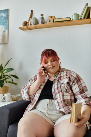 A woman sits on a couch, talking on a phone and holding a book.の写真素材