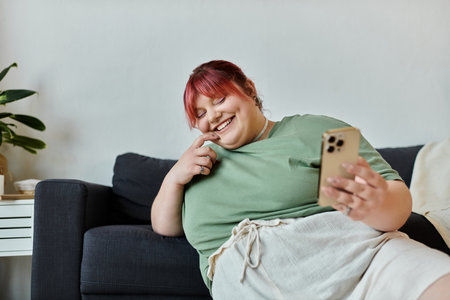 A woman laughs while sitting on a couch at home, using her phone.の写真素材