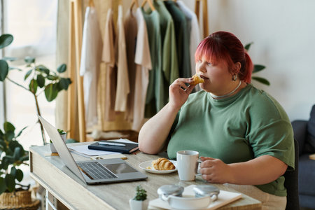 Red-haired woman eats pastry, drinks coffee, with open laptopの写真素材