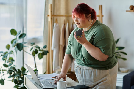 A woman stands at a table, laptop open, phone in hand, looking thoughtful.の写真素材