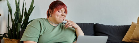 A plus size woman with pink hair sits on a couch in her home, smiling as she looks at a laptop.