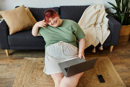 A woman sits on a rug, smiling as she uses a laptop.