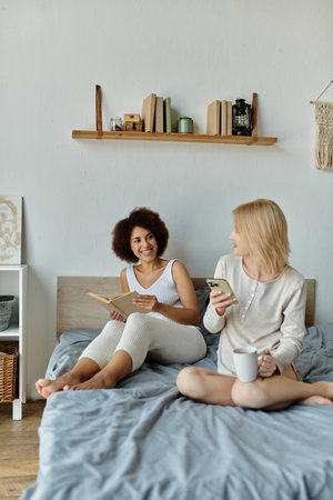 Two women, one reading a book and the other scrolling through her phone, relax together on a bed.の写真素材
