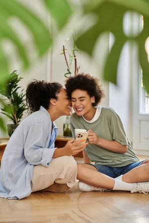 Two women sharing laughter while looking at a phone in their cozy living space.の写真素材