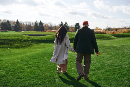 A loving couple dressed in cozy autumn attire strolls together across a lush green meadow.
