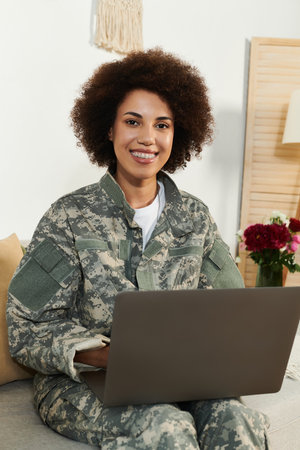 A confident young woman in camouflage uniform works on her laptop, smiling warmly in a cozy, bright setting.の写真素材