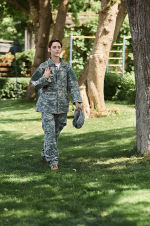A young soldier in camouflage uniform walks confidently through a green outdoor space, embracing...