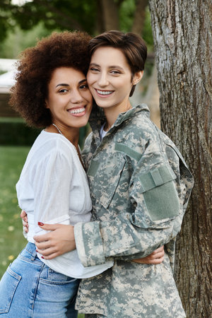 A female soldier in camouflage embraces her wife joyfully upon returning home in a serene park setting.の写真素材