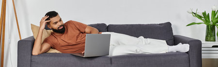 Handsome man lounges comfortably on the couch while browsing on his laptop.の写真素材