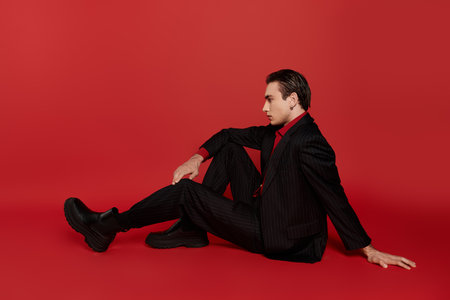 A man in a black suit and red shirt poses casually on a bright red backdrop.の写真素材