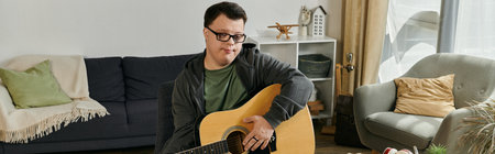 A young man with Down syndrome strums his guitar thoughtfully while sitting in a comfortable living...
