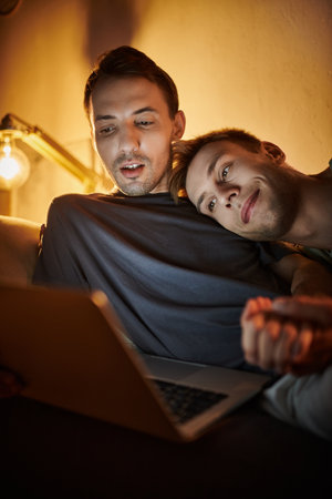 Two men cuddle together, sharing smiles while watching something on their laptop.の写真素材