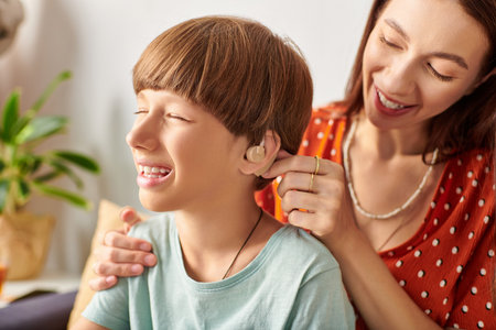 A mother lovingly assists her son with his hearing aid while smiling together.の写真素材
