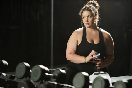 A plus-size woman lifts a water bottle, focused on her workout in a vibrant gym environment.