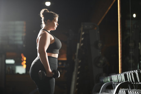 A young plus-size woman confidently lifts weights, promoting strength and body positivity in her...
