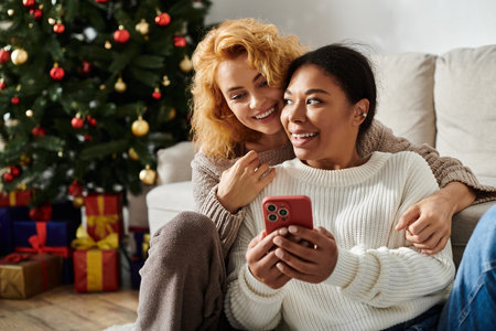 A couple enjoys cuddling together while looking at their phone, celebrating the holidays at home.の写真素材