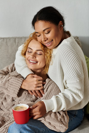 Two women embrace lovingly on a couch while savoring warm beverages in a cozy home setting.の写真素材