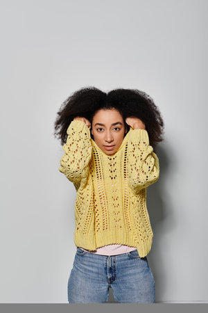 A young woman with curly hair showcases her style in a vibrant yellow sweater on a soft grey...