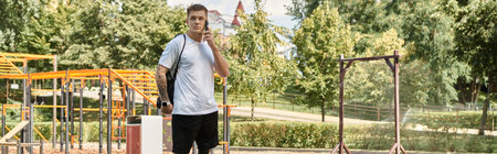 A young man with tattoos walks through a park, talking on his phone