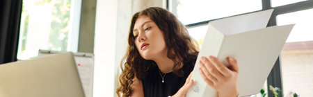 A young plus size woman with long curly hair focuses intently on her work at the office, surrounded...