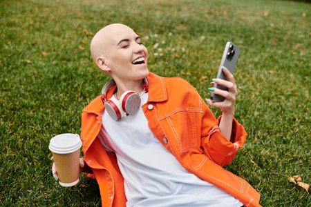 A joyful bald woman relaxes on the grass, sipping coffee and smiling at her phone.