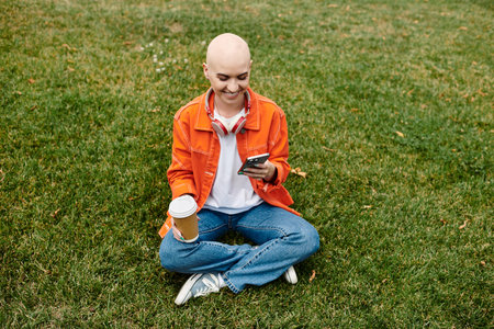 A young woman with a beautiful bald head smiles as she relaxes on the grass, holding coffee.の写真素材
