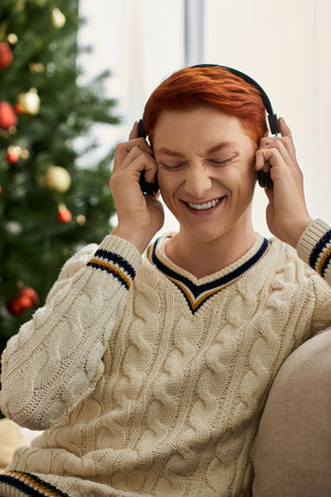 A delighted young man listens to music while seated beside a well decorated Christmas tree.