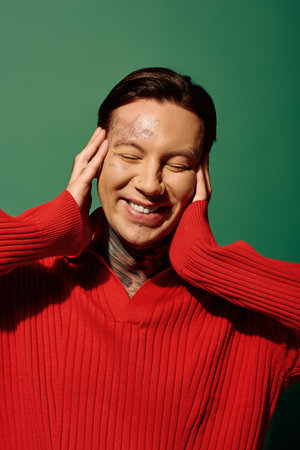 A young man with tattoos expresses joy while wearing a red sweater in a colorful background.