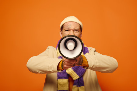 A young man with tattoos enthusiastically uses a megaphone against a bright orange backdrop.の写真素材