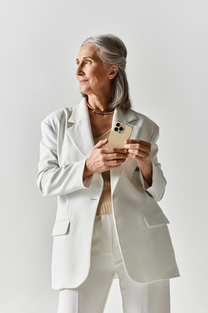 A fashionable mature woman wearing a white blazer and holding a smartphone showcases elegance and confidence.の写真素材