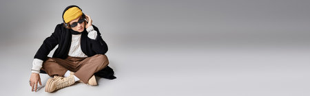 A handsome young man dressed in a black coat and yellow beanie lounges comfortably in a studio.