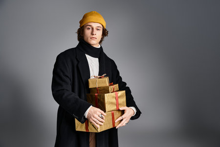 A young man stands confidently in a trendy black coat and yellow beanie, holding wrapped gifts in a studio.の写真素材