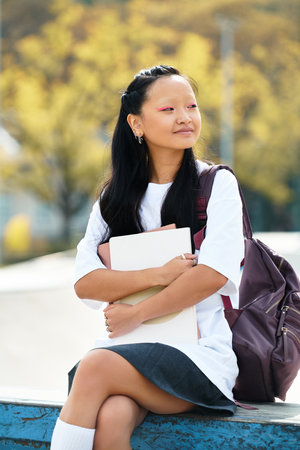 A stylish young Asian student relaxes outside, holding her books, embracing the spirit of youth and...